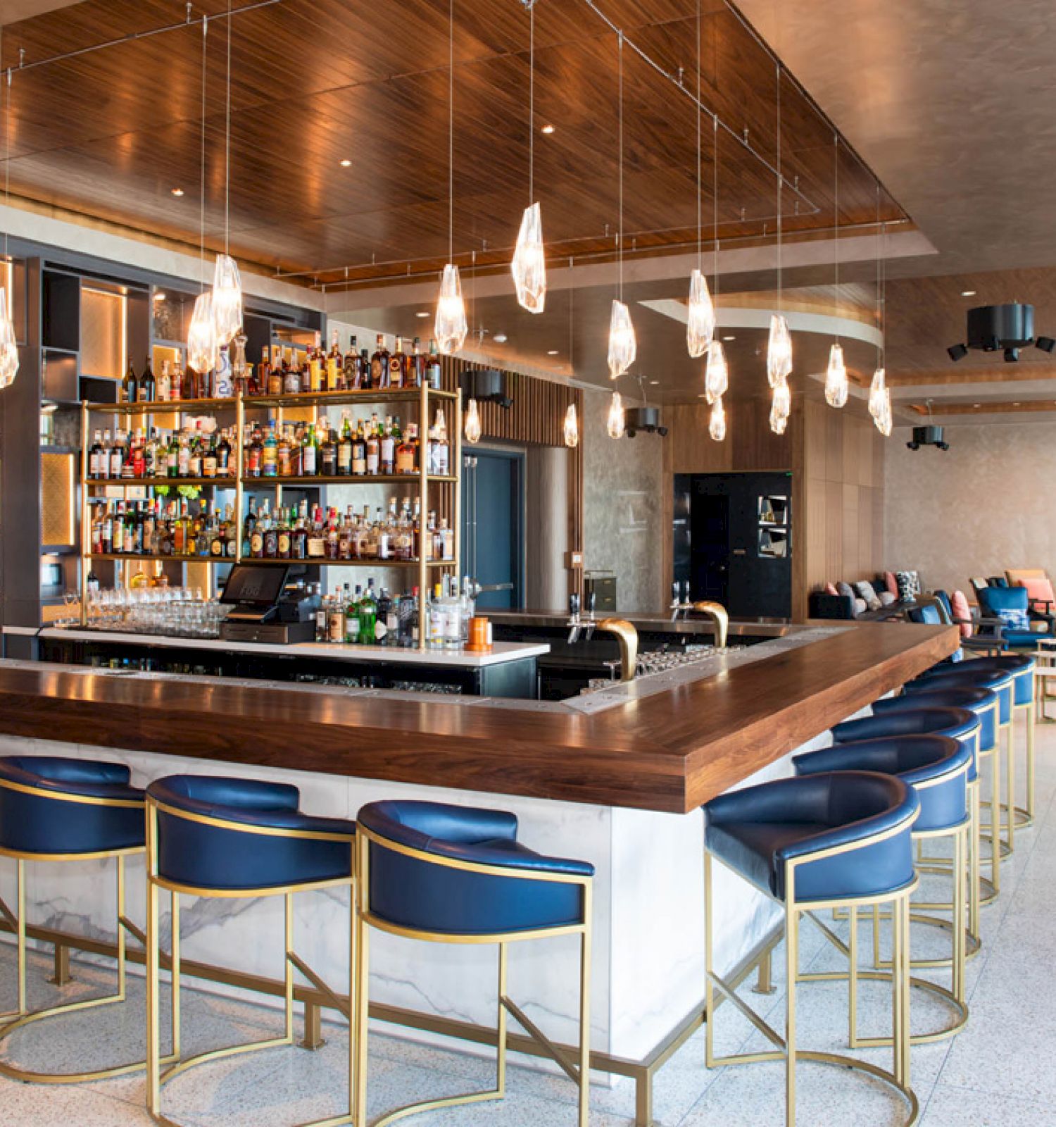 A modern bar with a wooden counter, blue stools, pendant lights, and a fully stocked back bar, set in a spacious, stylish lounge area.
