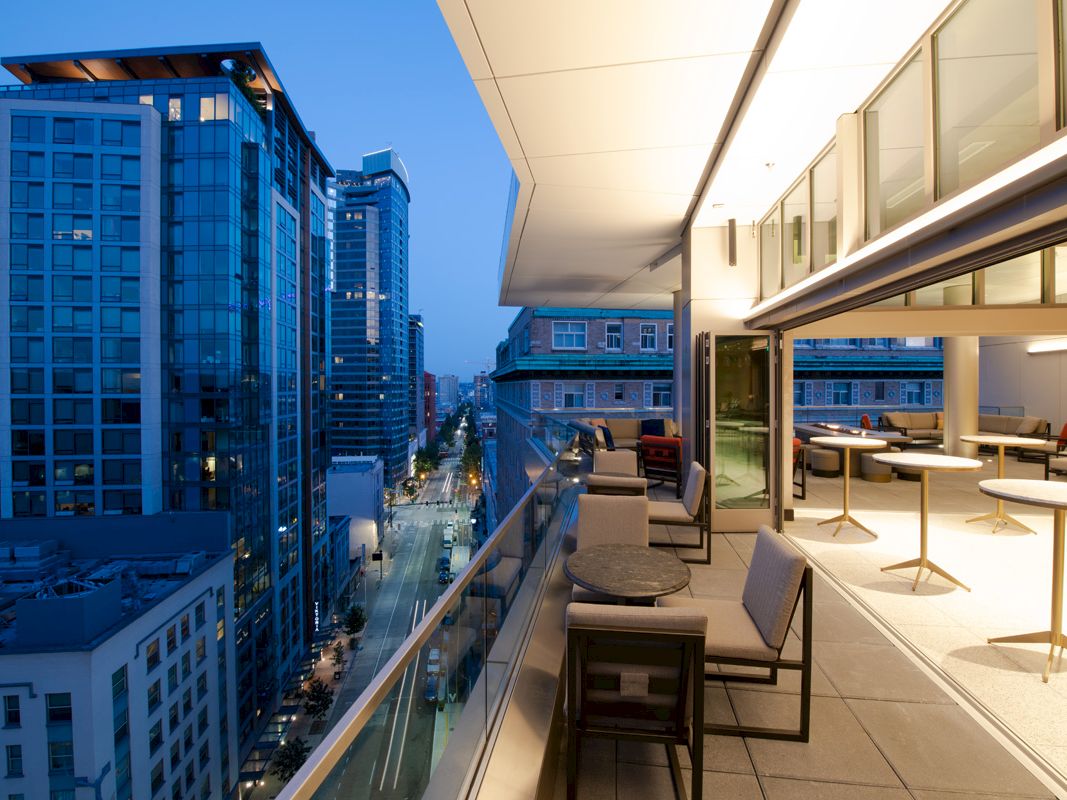 A modern cityscape at dusk with high-rise buildings, a lit balcony with tables and chairs, and a view of the street below.
