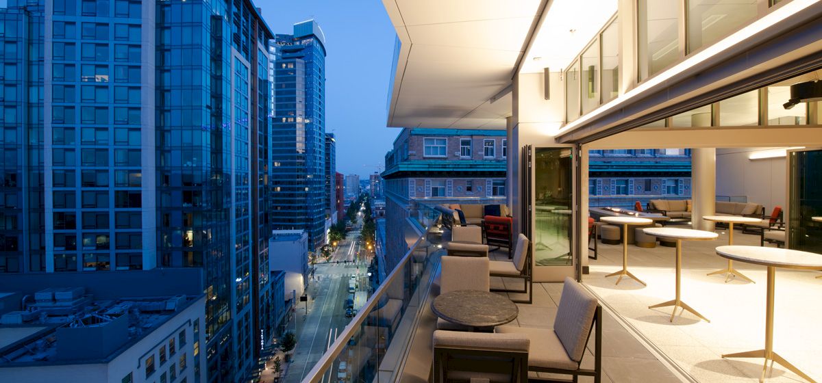 A modern cityscape at dusk with high-rise buildings, a lit balcony with tables and chairs, and a view of the street below.