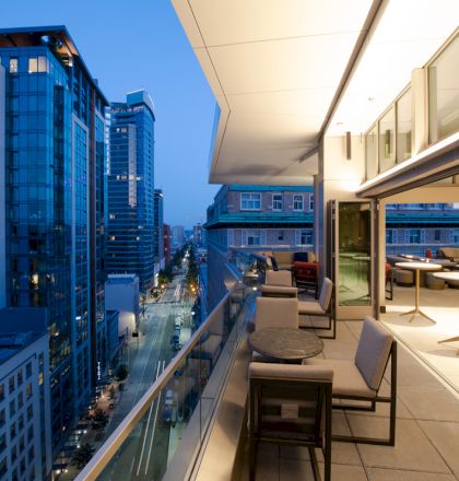 The image shows a modern cityscape with tall buildings and a balcony featuring tables and chairs, under soft evening lighting.