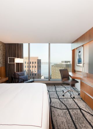 A modern hotel room with a large bed, TV, desk, and a window with a city view is shown in the image.