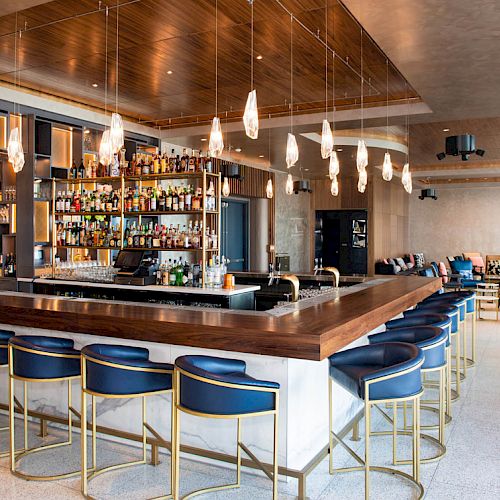 A modern bar with a wooden counter, surrounded by blue stools, overhead lights, and shelves stocked with an array of bottles and glasses.