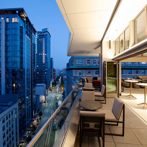 A modern balcony with seating overlooks a cityscape of tall buildings at dusk, with indoor areas visible through a glass wall.