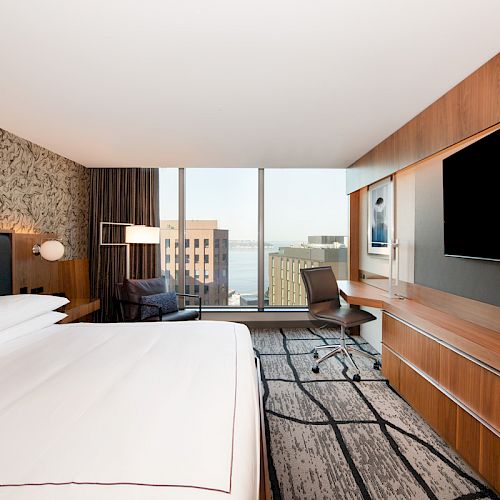 A modern hotel room with a large bed, desk, chair, and wall-mounted TV. The window offers a city and water view.