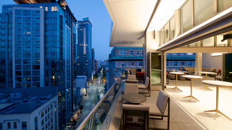 A modern cityscape with tall buildings, a sleek balcony with seating, and warm lighting during evening hours.