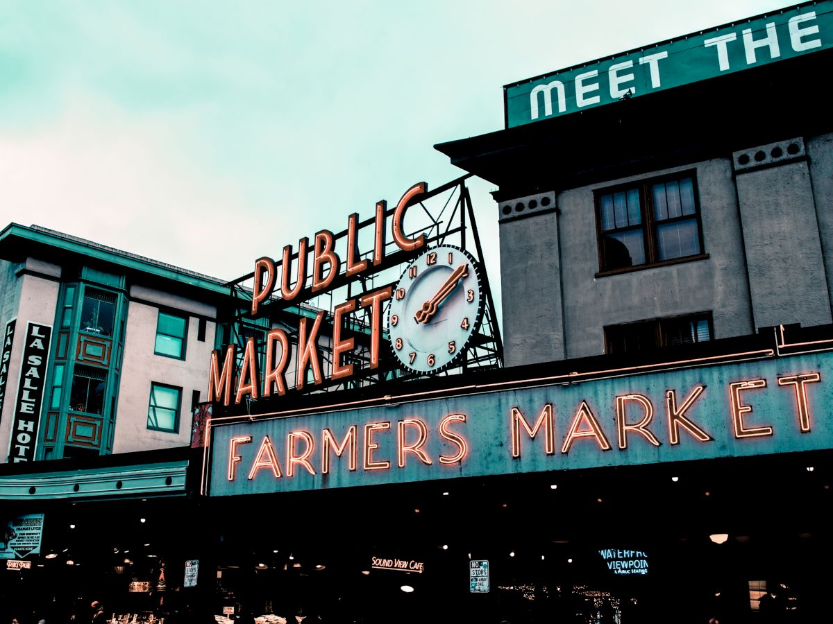 The image shows the famous neon signs for "Public Market" and "Farmers Market" at Pike Place Market in Seattle, with buildings in the background.