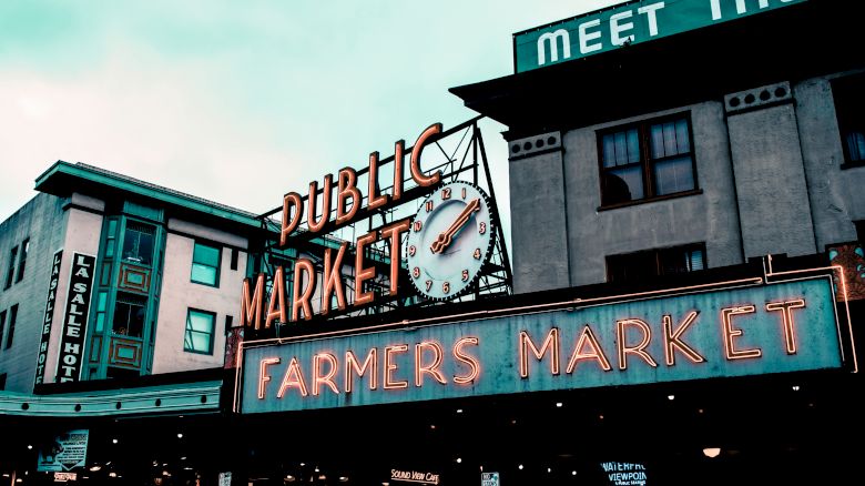 The image shows the famous neon signs for "Public Market" and "Farmers Market" at Pike Place Market in Seattle, with buildings in the background.