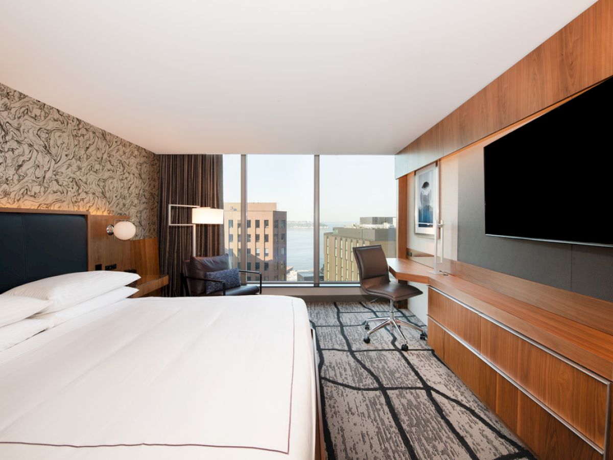 A modern hotel room with a large bed, TV, desk, chair, and window view of buildings and water.