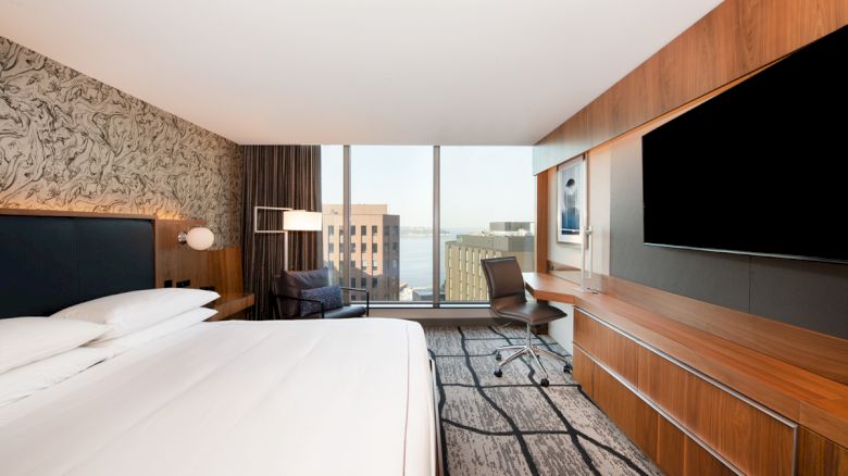A modern hotel room with a large bed, TV, desk, chair, and window view of buildings and water.