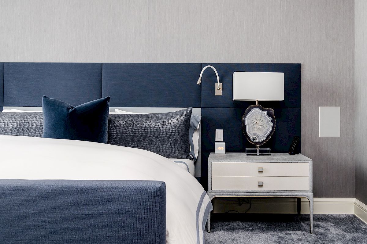 A modern bedroom with a blue bed, two pillows, a nightstand with a lamp, and decorative items. The room features a minimalist design.