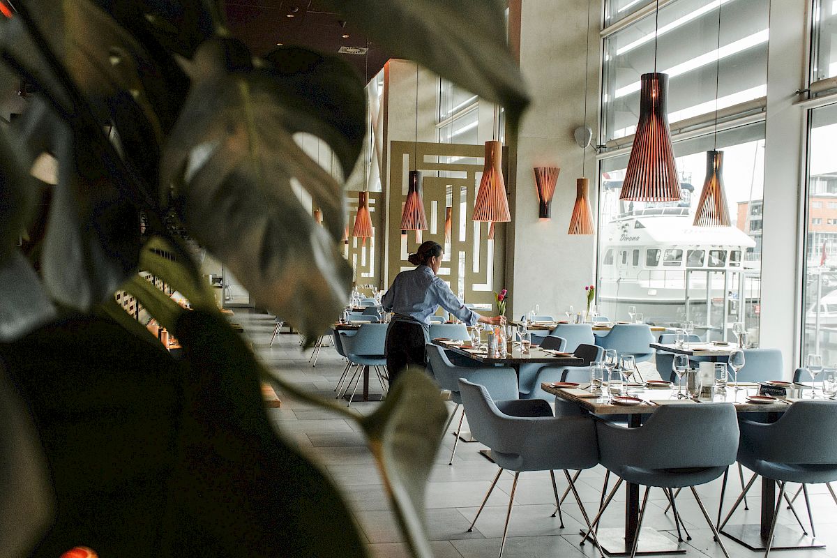 A modern restaurant interior with stylish pendant lamps, elegant table settings, and a person arranging a table, viewed through green leaves.